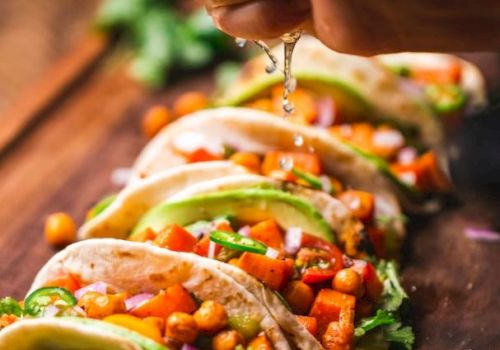 The image shows several tacos filled with chickpeas, vegetables, and herbs on a wooden surface, with someone squeezing lime over them.