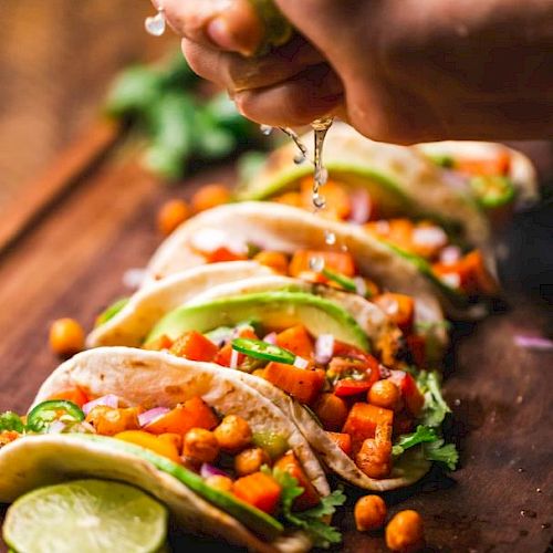 The image shows several tacos filled with chickpeas, vegetables, and herbs on a wooden surface, with someone squeezing lime over them.