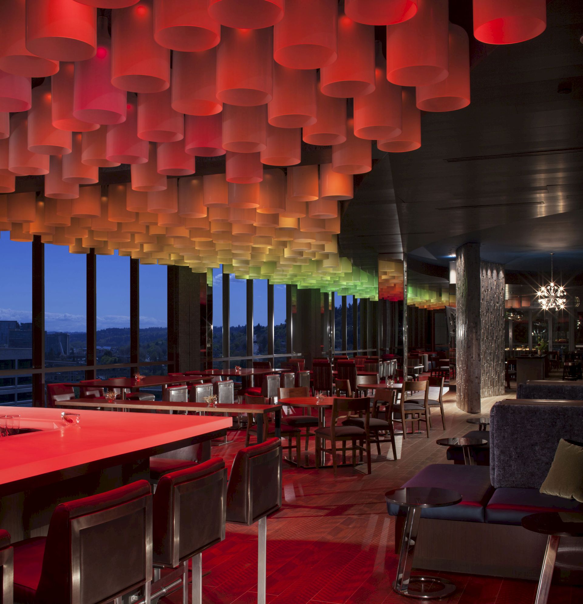 The image shows a modern, stylish bar or restaurant interior with colorful ceiling lights, high tables, and chairs, and large windows with a nighttime view.