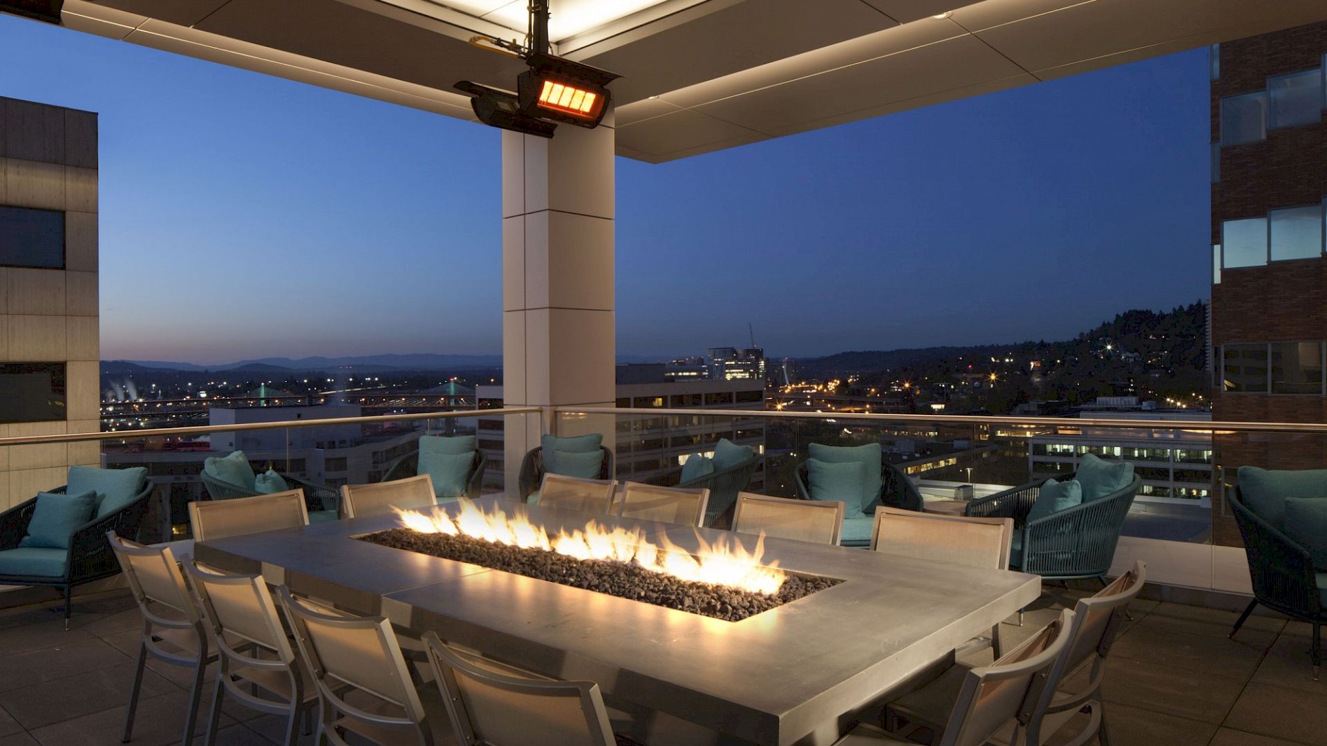 The image shows a rooftop patio with a long rectangular fire table, surrounded by chairs, overlooking a cityscape at dusk with buildings and hills in the background.