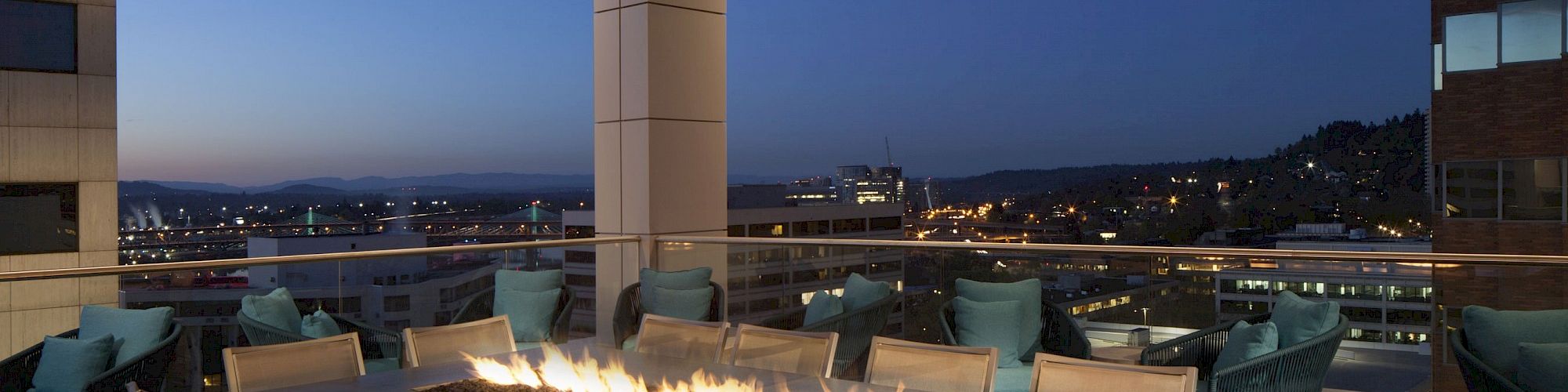 The image shows a rooftop patio with a long rectangular fire table, surrounded by chairs, overlooking a cityscape at dusk with buildings and hills in the background.