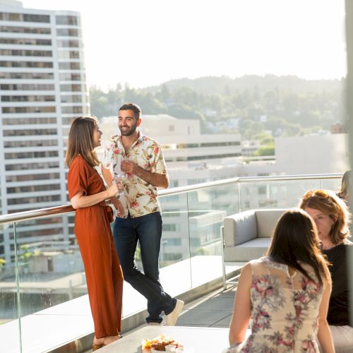 A group of people socializing on a rooftop terrace, with some standing by the railing and others sitting at a table against a city backdrop.