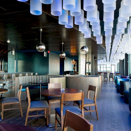 A modern bar with wooden tables and chairs, an illuminated cylindrical ceiling design, blue-lit ambiance, and a long counter.