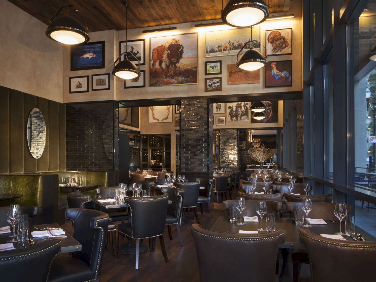 This image shows a cozy, modern restaurant with wooden decor, framed artwork, leather chairs, and tables set with wine glasses and napkins, ready for diners.