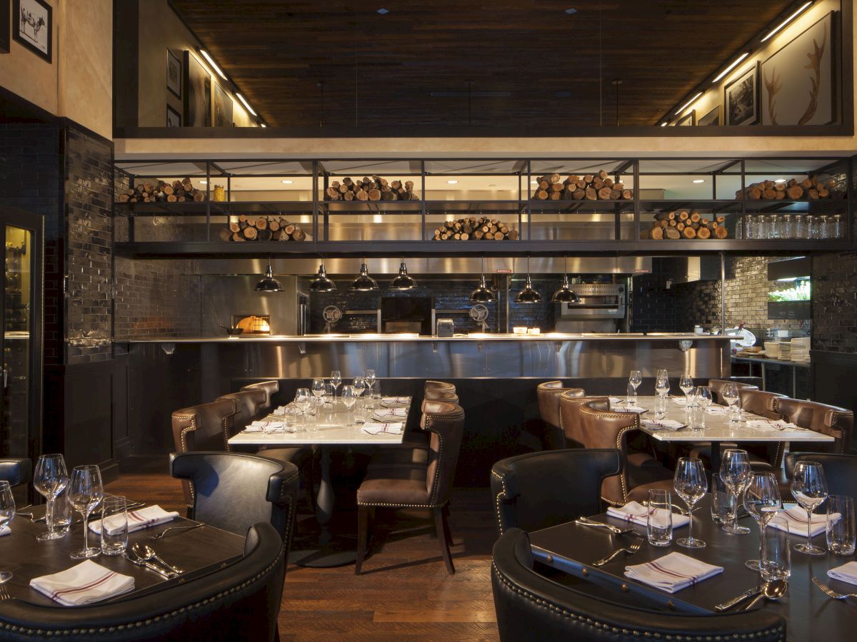 A modern restaurant interior with neatly set tables, leather chairs, wine glasses, and an open kitchen with stacked firewood and warm lighting.