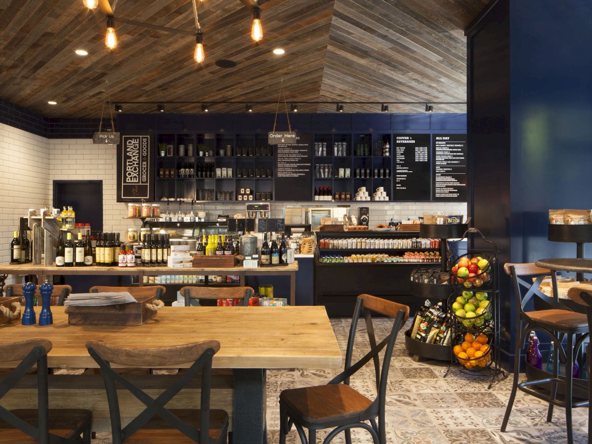 A cozy café interior with wooden tables, chairs, and a counter displaying various items for sale. Fruits are arranged in a tiered stand.