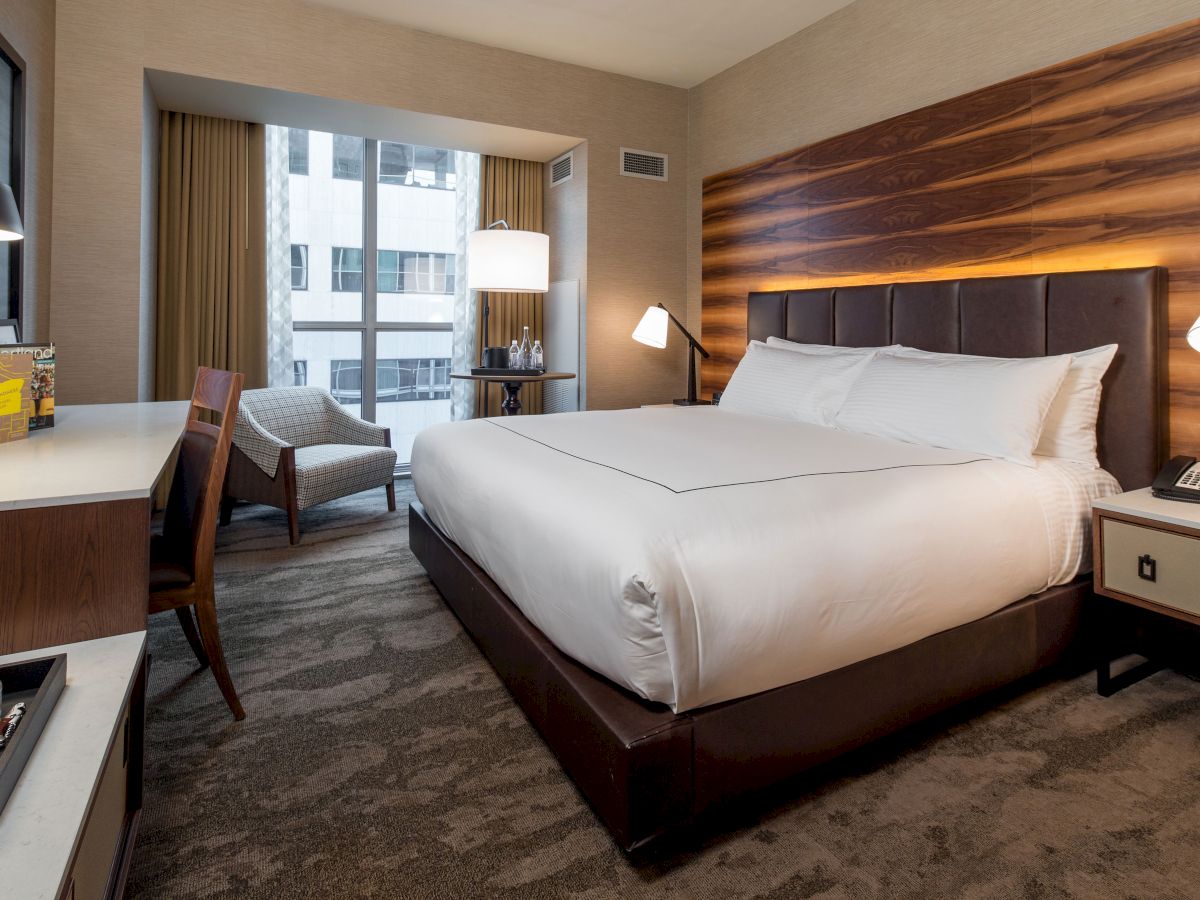 This image shows a hotel room with a neatly made bed, a desk and chair, a lounge chair by the window, and modern decor, providing a cozy ambiance.