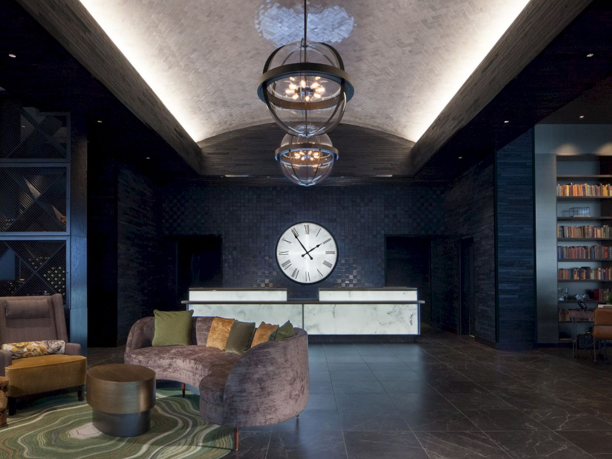 The image shows a stylish lobby with a large wall clock, modern lighting, a curved sofa with cushions, and a bookshelf in a cozy, contemporary setting.