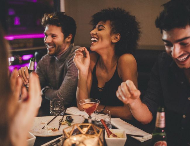 A group of friends is enjoying a night out together, laughing and having drinks at a dimly lit restaurant or bar, creating a lively atmosphere.