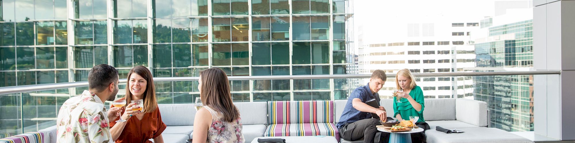 People are enjoying drinks and food on a rooftop patio with modern seating and a fire pit, surrounded by city buildings.